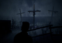 A hooded man looking out at crucifixes in a field