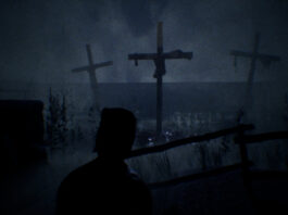 A hooded man looking out at crucifixes in a field