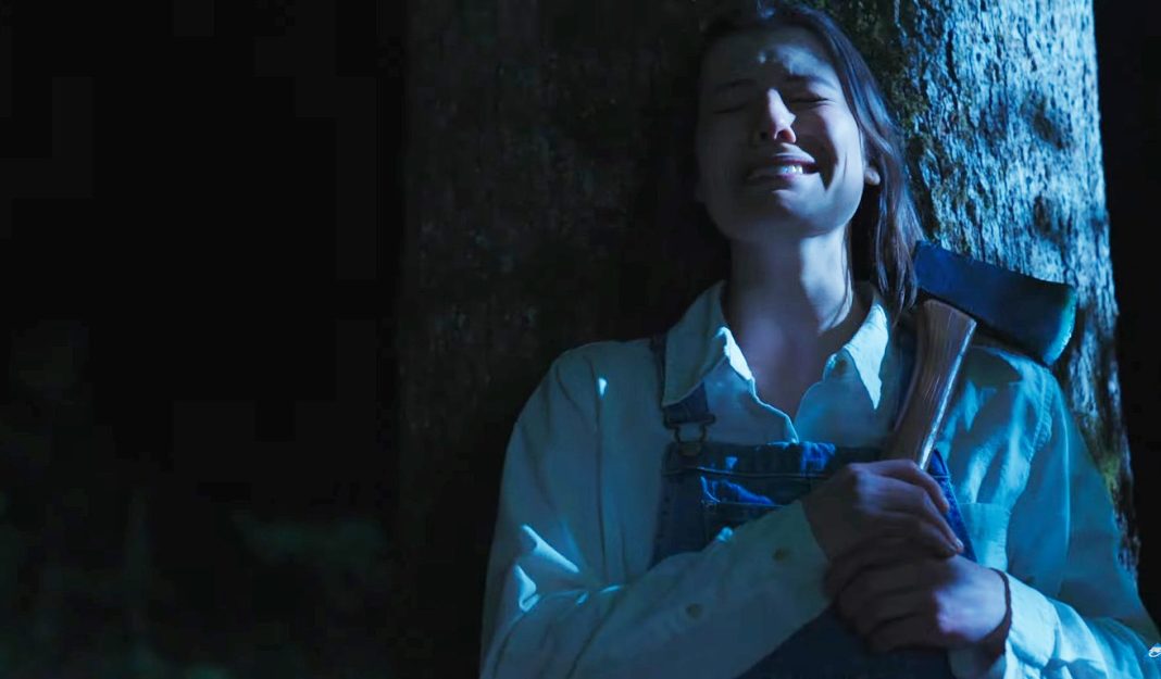 A woman stands against a tree a night, holding an axe, while crying