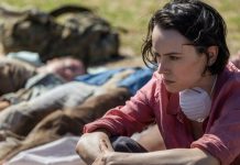 ‘We Bury The Dead’ Review: 2026’s First Horror Movie Puts a New Spin on the Zombie Subgenre A woman sits next to dead bodies while wearing a mask around her neck in We Bury the Dead