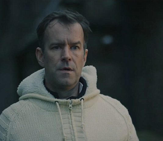 A man wearing a white sweater stares at someone in a graveyard in Watch Me Sleep