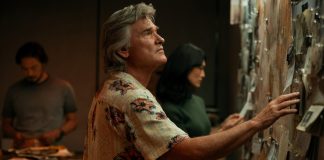 Wearing a floral shirt, Lee Shaw (Kurt Russell) looks at a wall covered in documents in Monarch Legacy of Monsters Season 2, Episode 4 'Trespass'