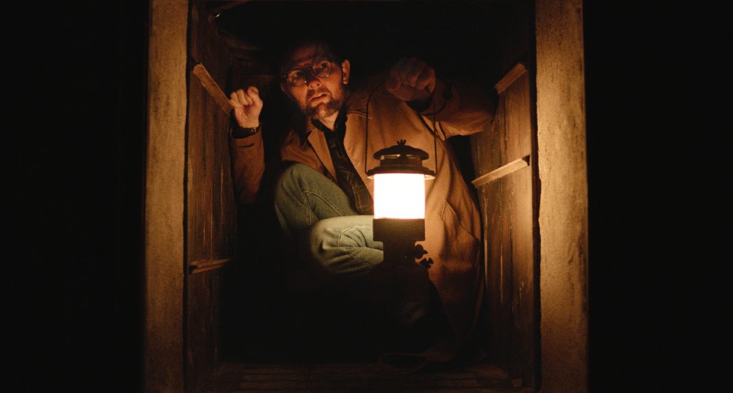 Adam Scott's character hides in a crawl space with a lantern in Hokum