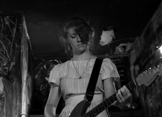 A woman plays the guitar while on stage in The Yellow Bellies