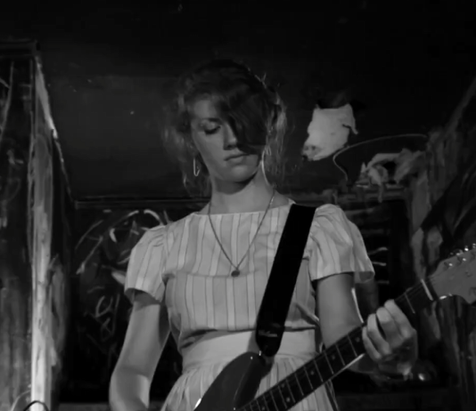 A woman plays the guitar while on stage in The Yellow Bellies