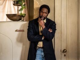 Boyd stands in the kitchen next to the fridge, thinking, in FROM Season 4 Episode 6