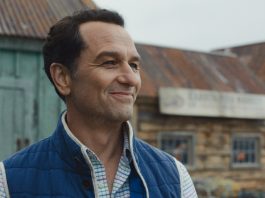 Tom Loftis smiles as he looks around outside in Widows Bay Season 1
