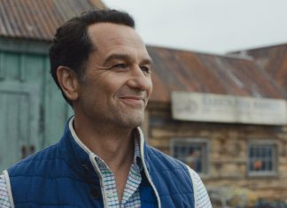 Tom Loftis smiles as he looks around outside in Widows Bay Season 1