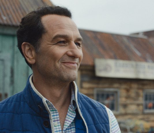 Tom Loftis smiles as he looks around outside in Widows Bay Season 1